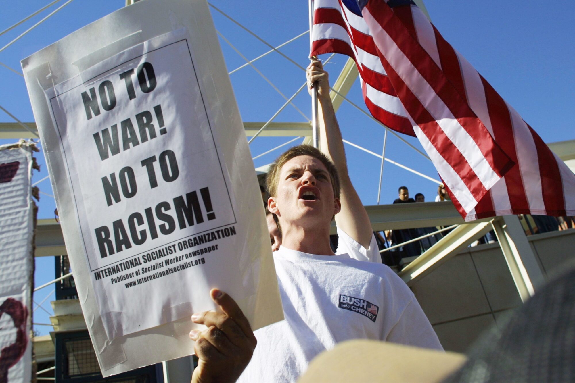Protestor holding a sign that says "No to war! No to Racism!" with American flag in background