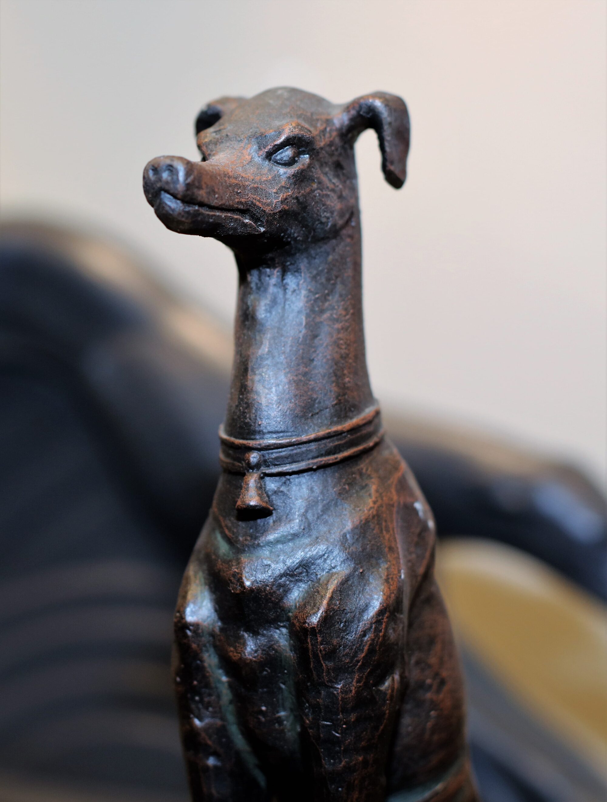 Sculpture of a sitting greyhound in semi-profile with a bell on its collar.