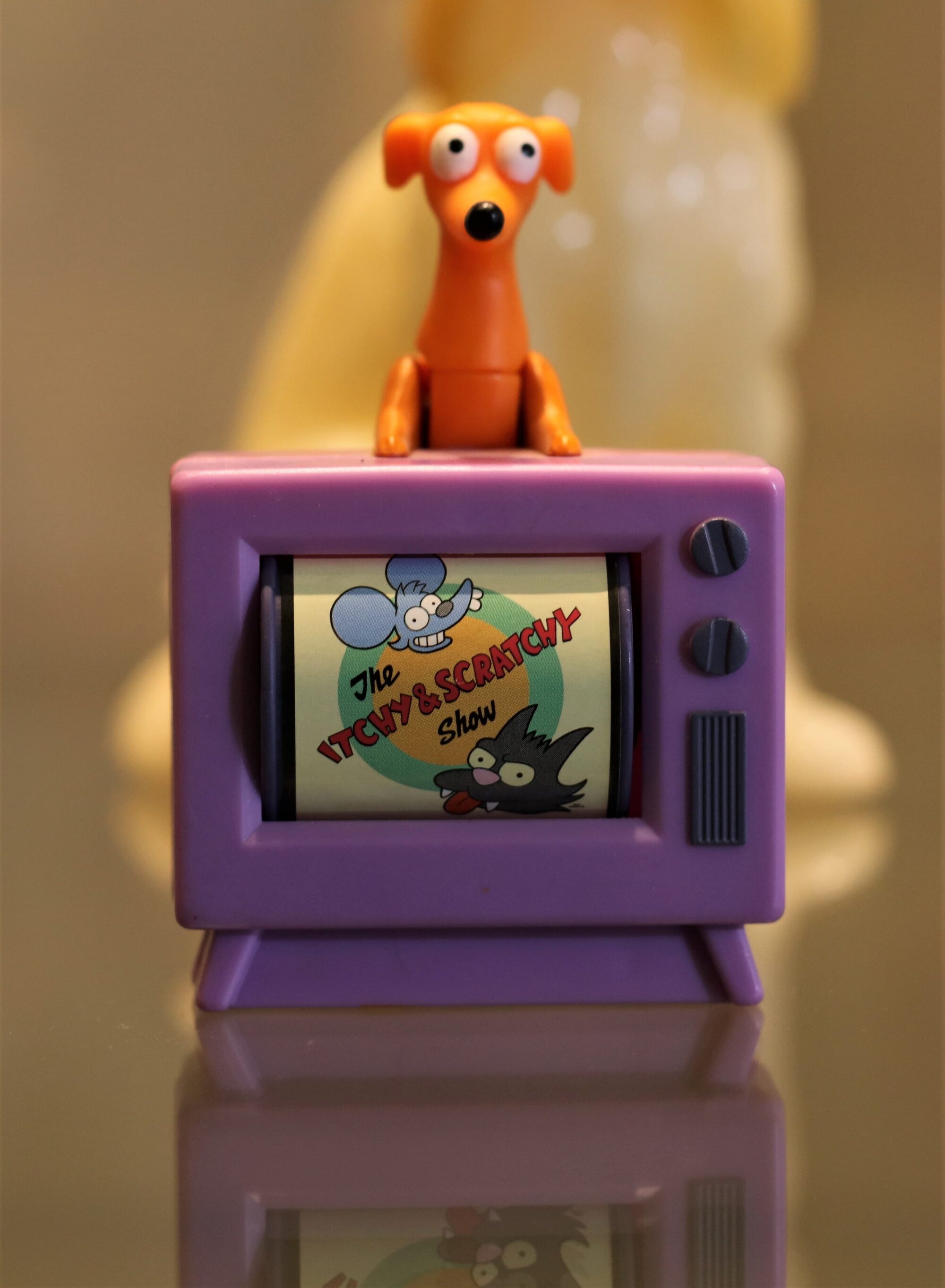 A small plastic toy consisting of a purple TV set with a flip screen showing a mouse and a cat and text reading The Itchy and Scratchy Show, with an orange greyhound standing behind the set.