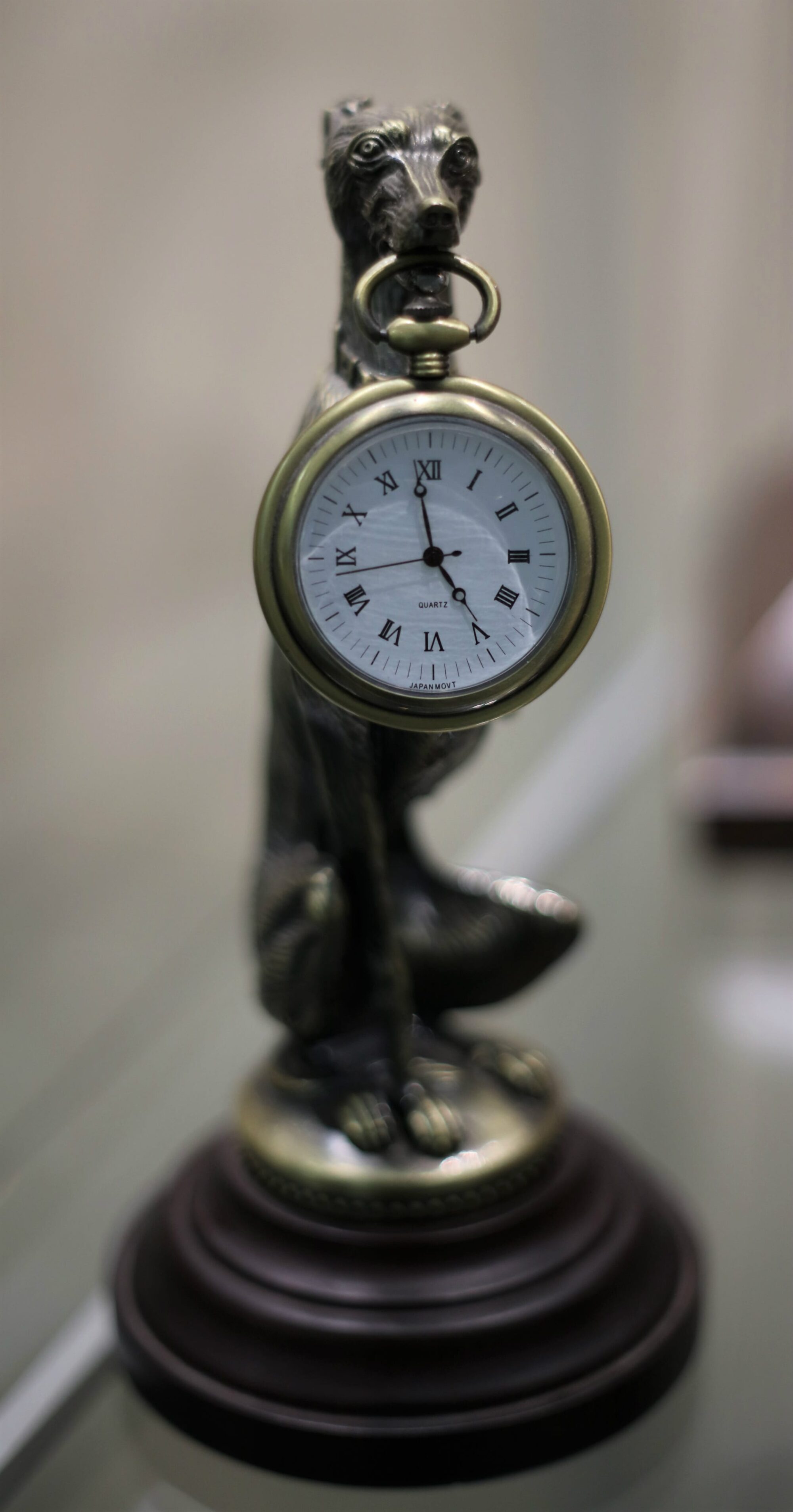 Sculpture of a greyhound holding a small clock in its mouth on a pedestal.