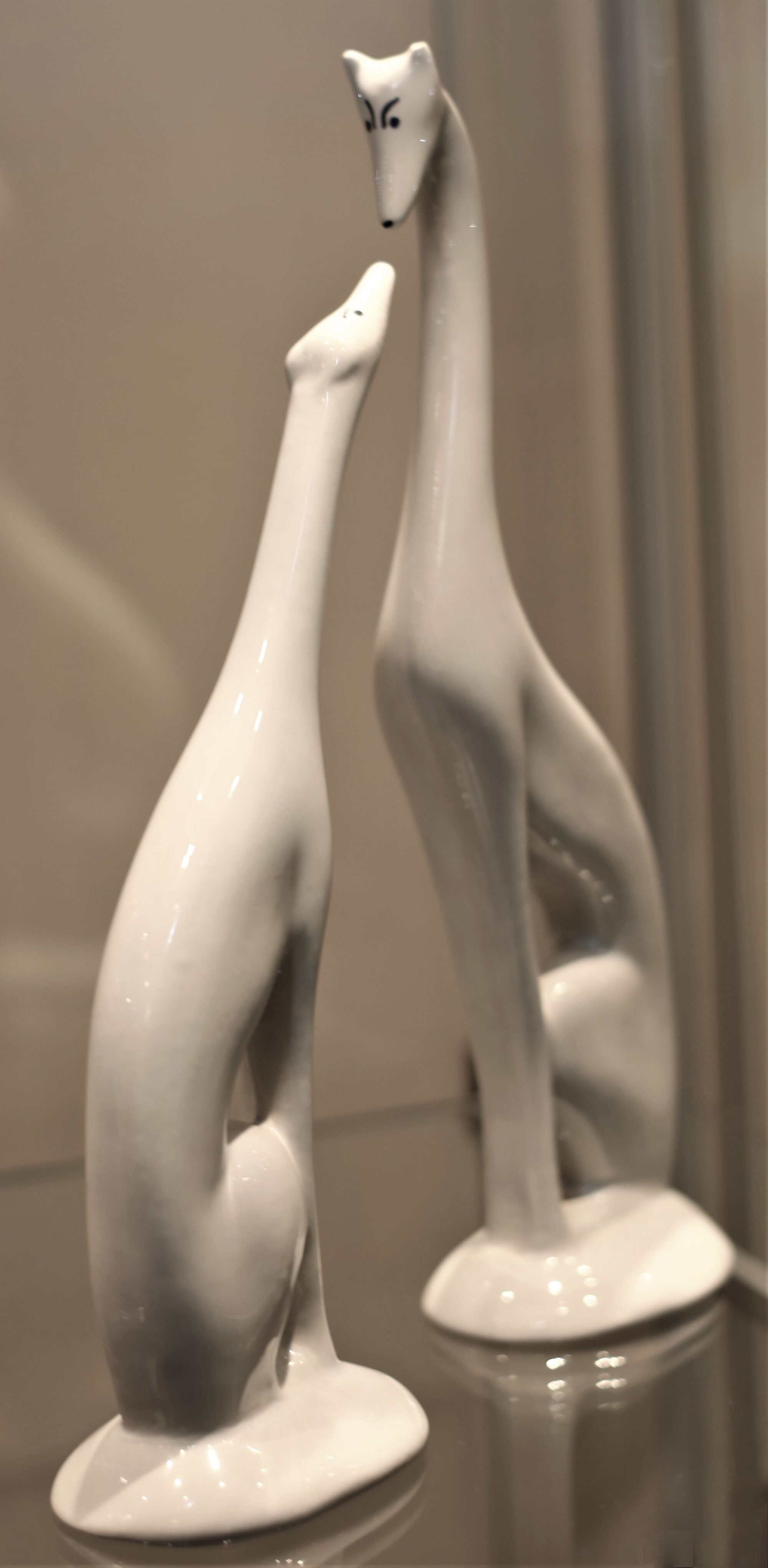Two ceramics of slightly abstract white greyhounds nose to nose. Both are sitting, but one is haunched.