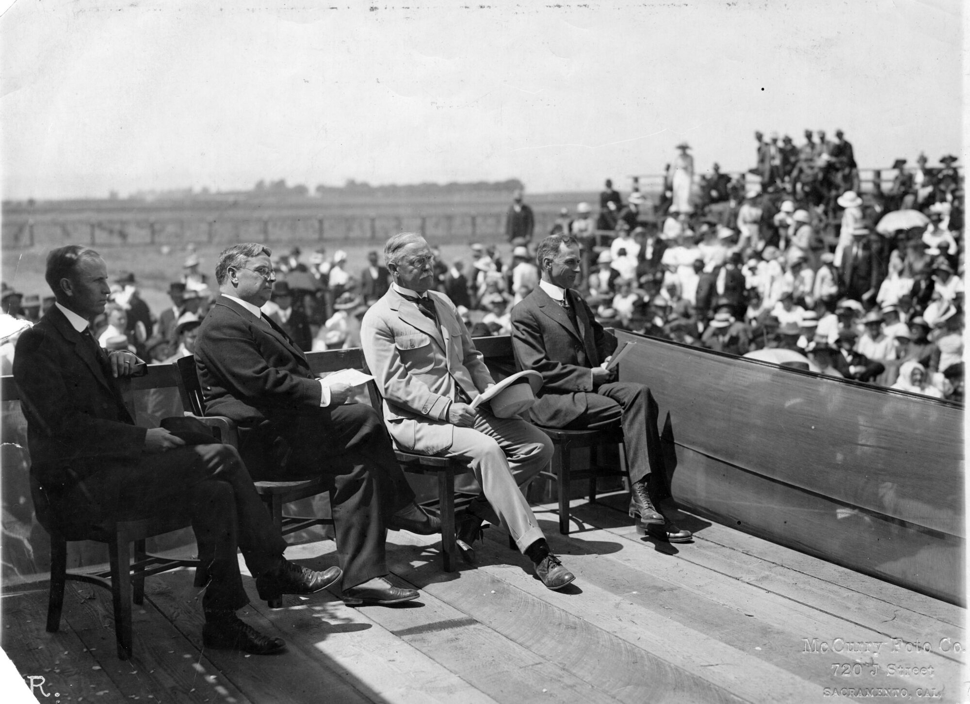 Four men in suits sitting in chairs on a raised platform, blurred crowd in the background