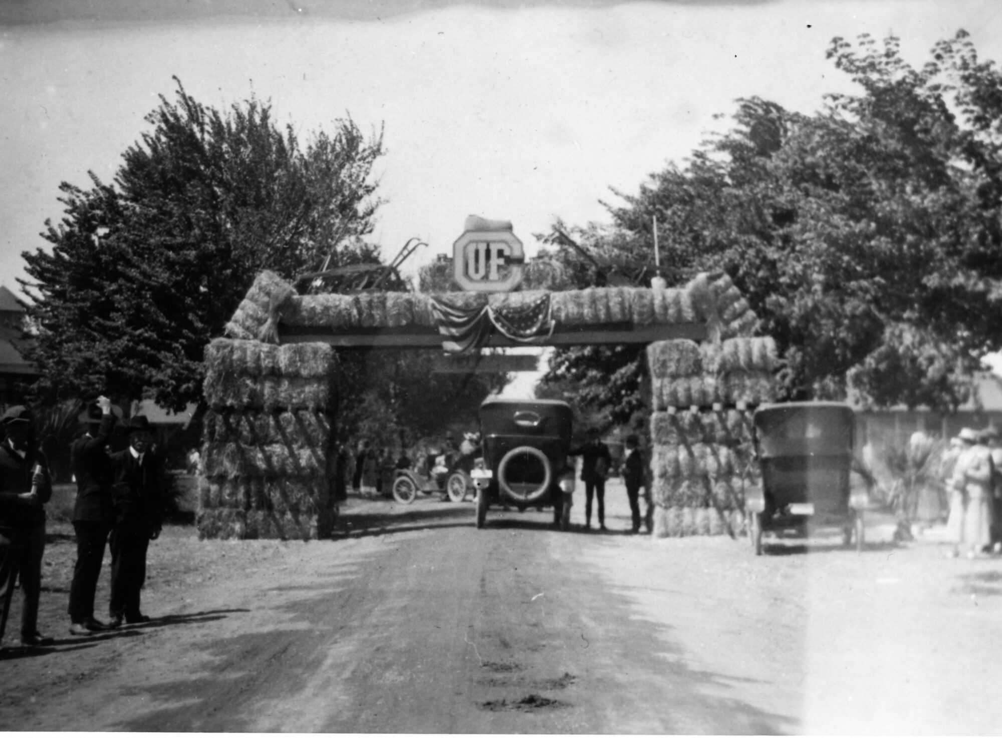 Car passing under an arch of stacked hay bales with a draped US  flag and the initials UF.