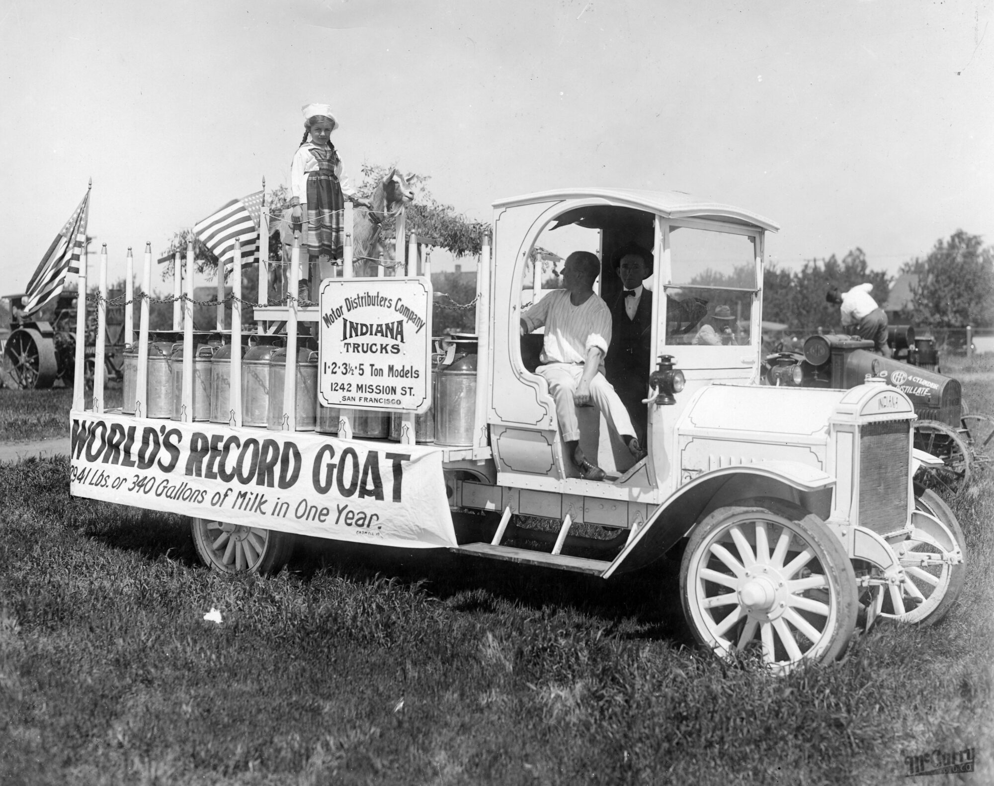 Small girl and goat standing on the back of a milk truck with American flags and a sign that says "World's Record Goat: 2941 lbs or 340 gallons of milk in one year"