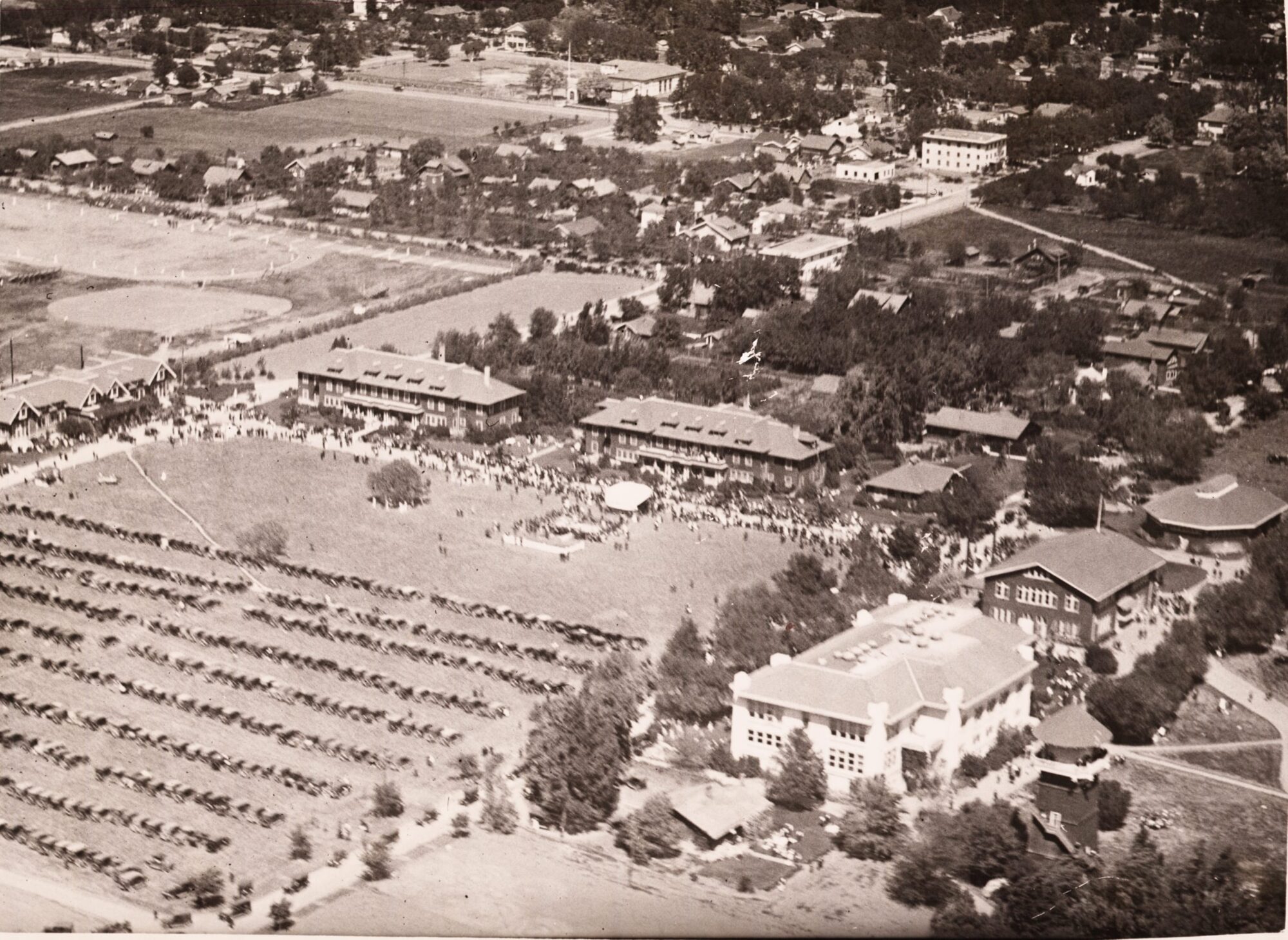 Aerial view of campus with many people lining the streets and rows of parked cars in a nearby field