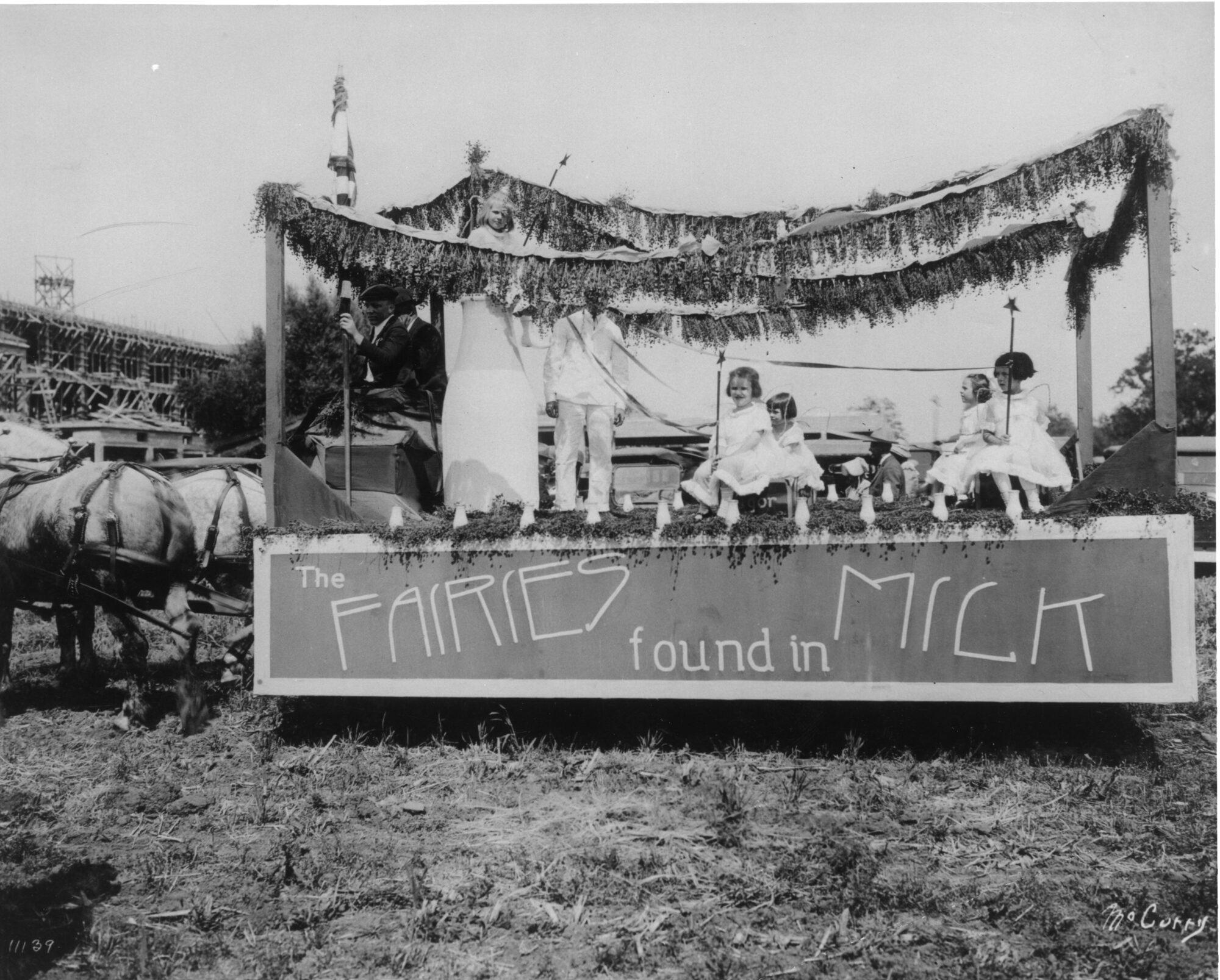 Little girls sitting on a horse-drawn wagon float draped with foliage and a giant milk bottle labeled: the fairies found in milk