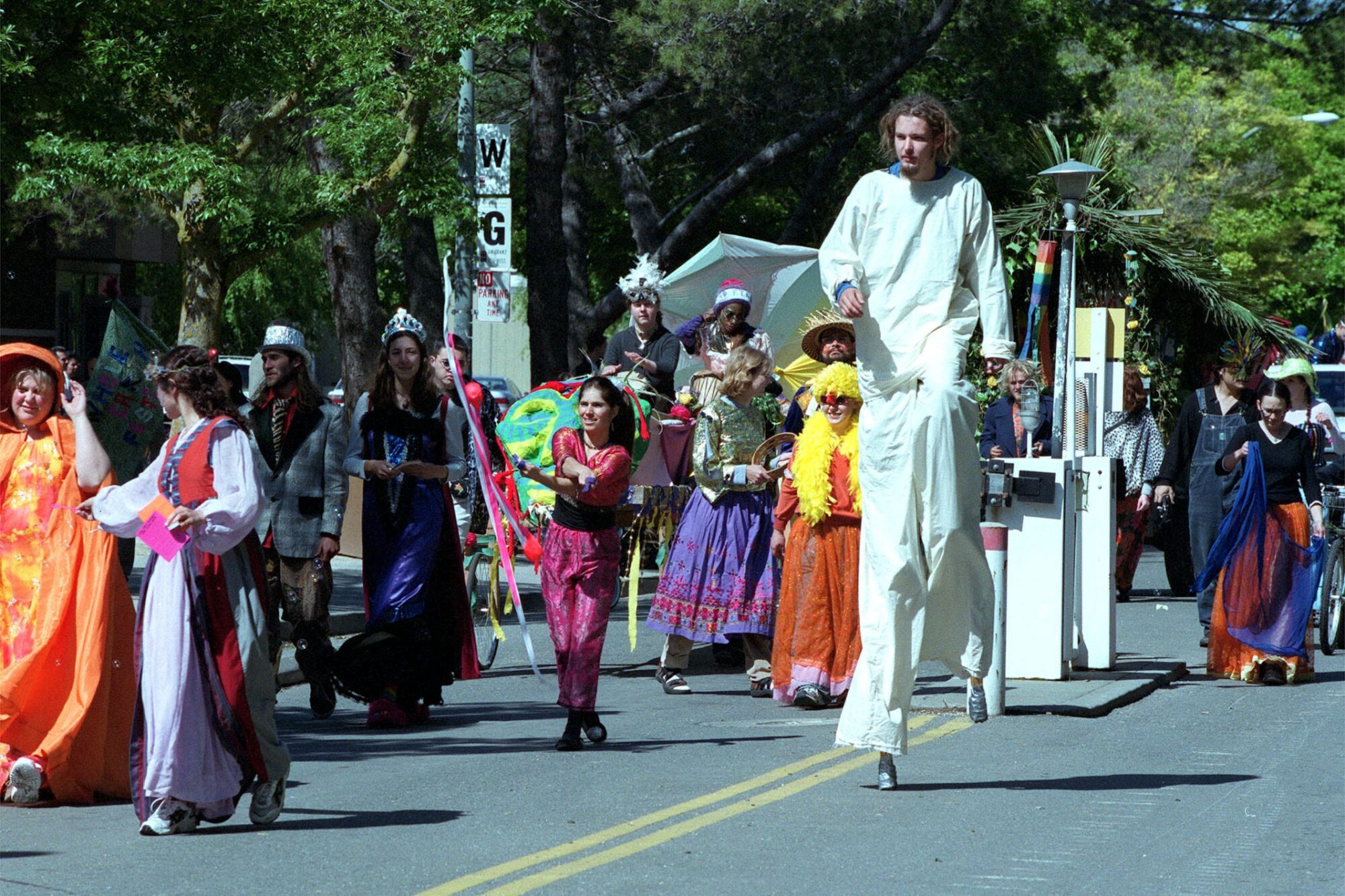 A group of people in colorful costumes, including a person on tall stilts, walking in the Picnic Day parade