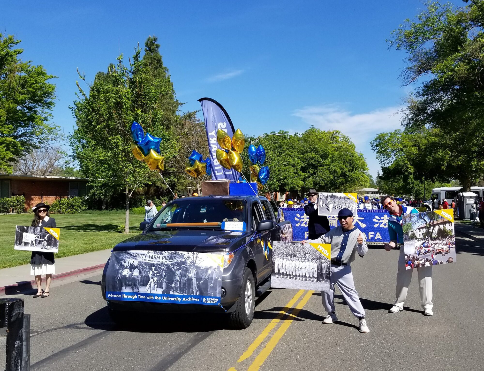 Library employees around a balloon-decorated truck dressed in period attire holding images from matching decades: 1910s, 1920, 1940s, and 1980s
