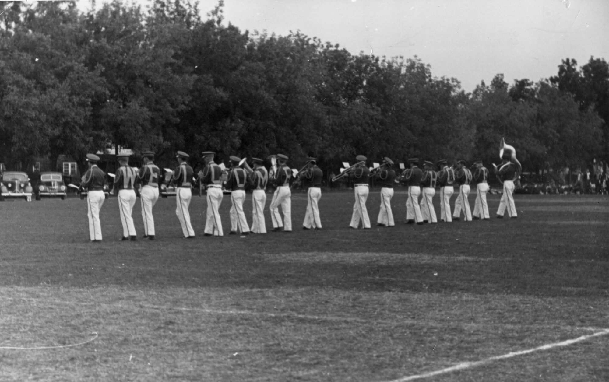 The backs of the marching band playing instruments in uniform with each player wearing a letter to spell "welcome home"