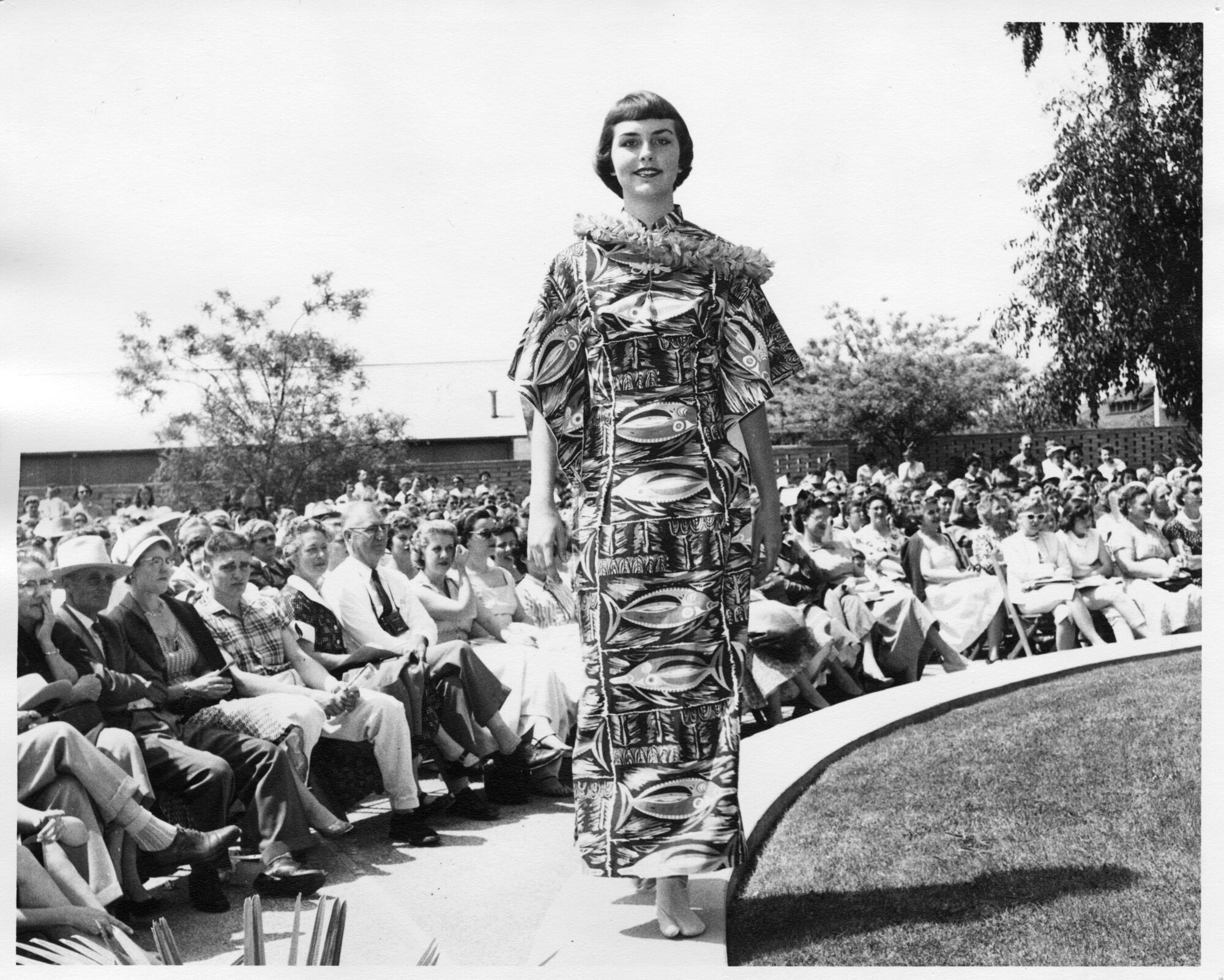 Woman with short hair wearing a wide sleeved fish printed dress and flower lei on a runway