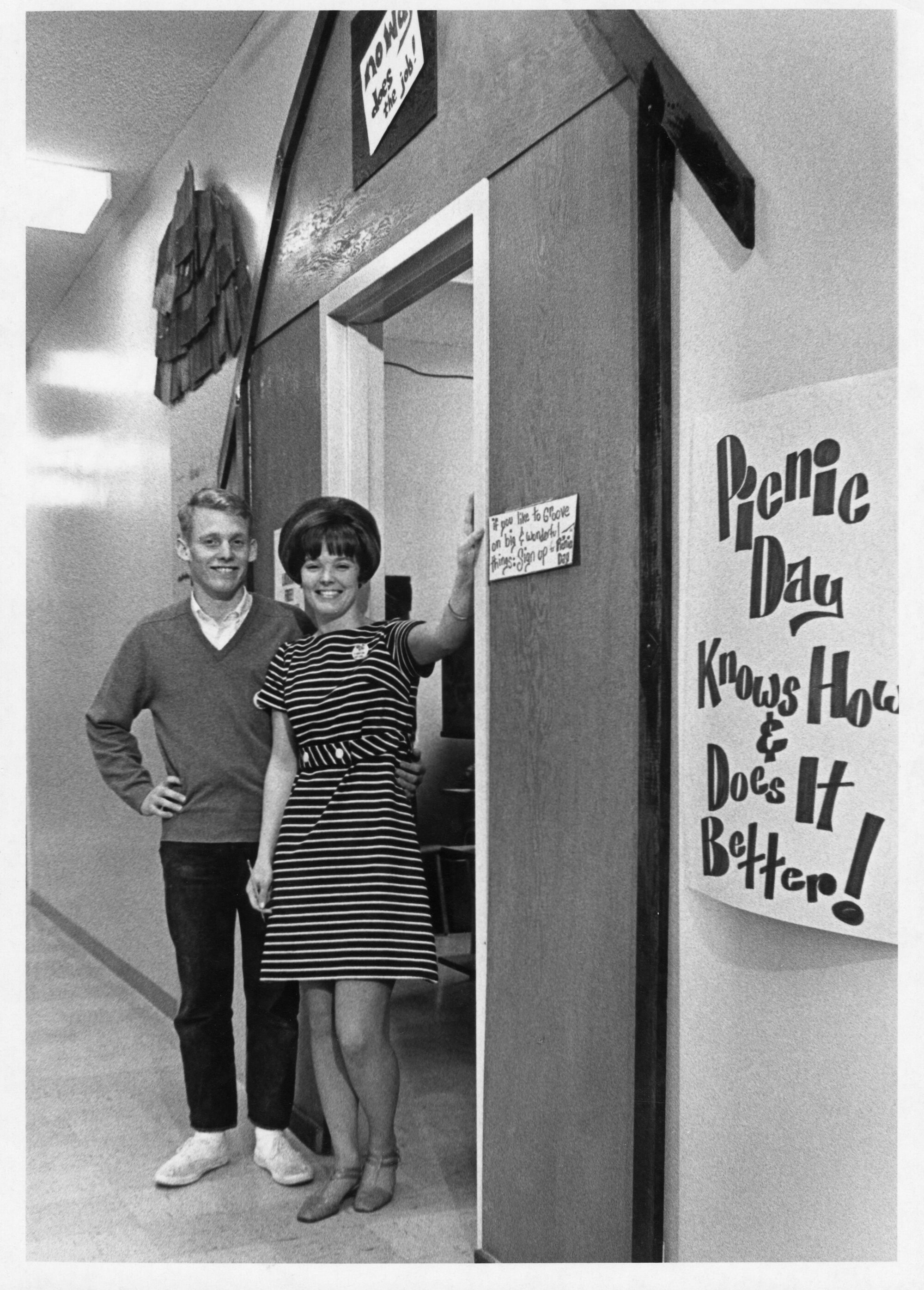 Picnic Day Chairman and Hostess pose in a mock barn doorway beside a large poster reading "Picnic Day Knows How & Does It Better!", 1968