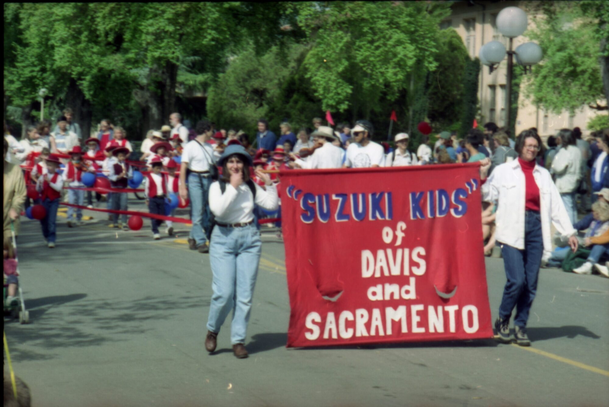 Kids in red vests led by a pair of adults holding a sign that says Suzuki kids of Davis and Sacramento
