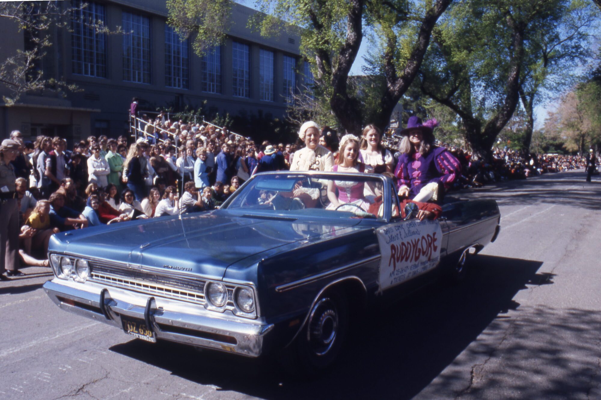 Students in colorful historical garb riding in the back of an open top convertible Plymouth car
