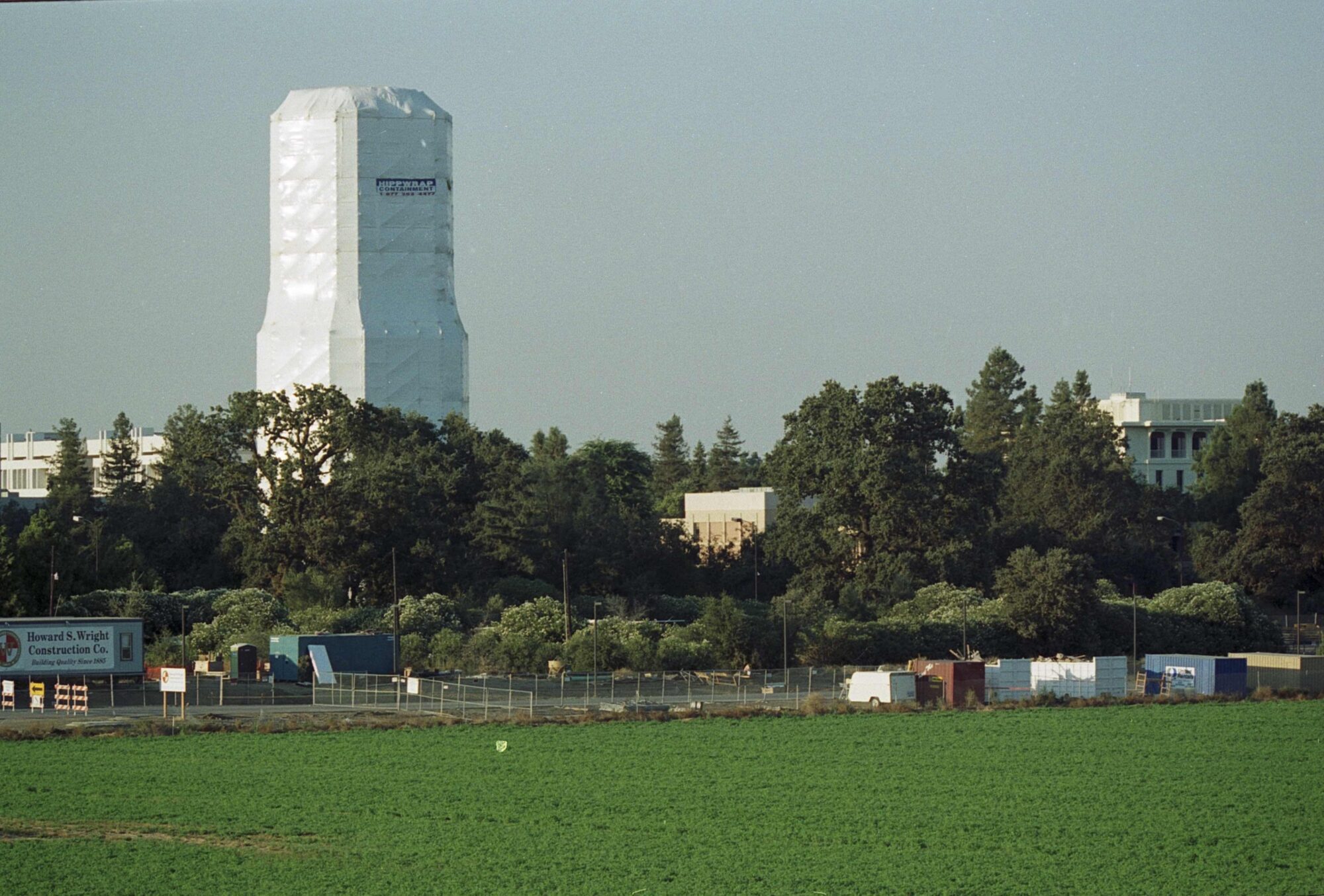 Water tower under white covering for renovation viewed behind tree line. 