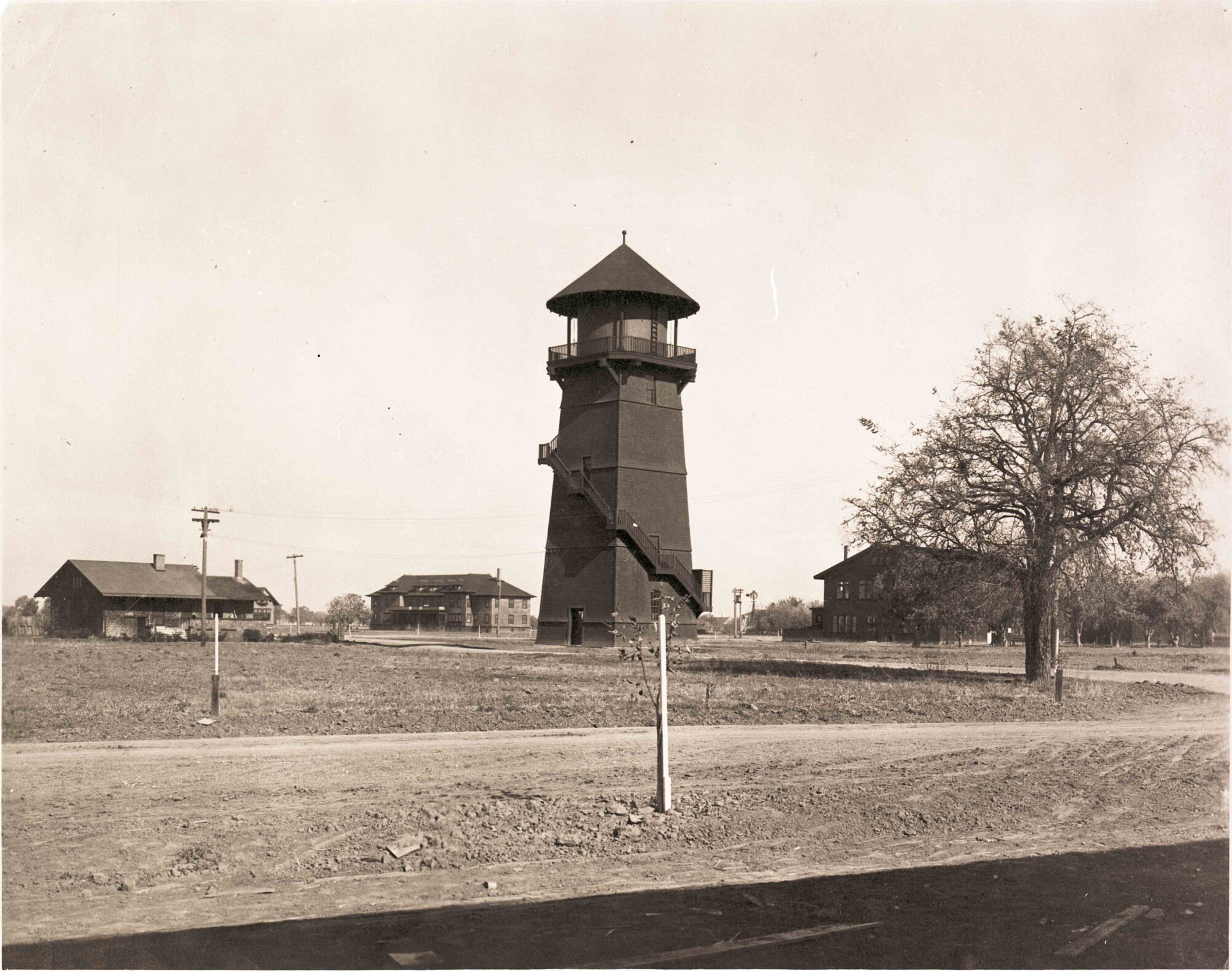 A black and white, sepia toned photograph of a stout, wooden-framed water tower with a conical top surrounded by sparse trees and the early campus buildings. 