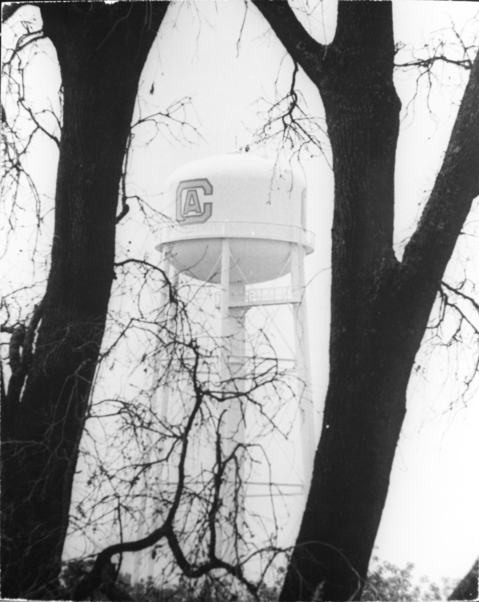 Black and white photograph of water tower with CA painted on it viewed through trees