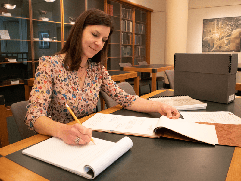 A woman sitting at a green desk with a document case beside her taking notes on some of the materials.