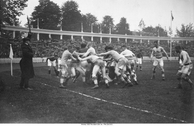 A black and white photograph of a rugby game. A group of men are in the midst of tackling each other.