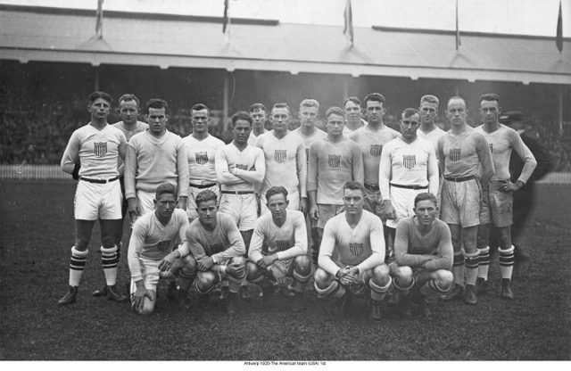 A black and white photograph of a group of men in rugby uniforms posing on a rugby film with a large audience behind them.