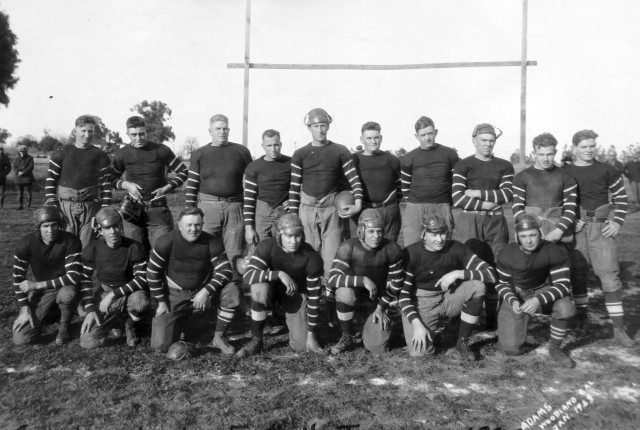 A black and white photograph of a group of young men in rugby gear and uniforms posing outside on a rugby field.