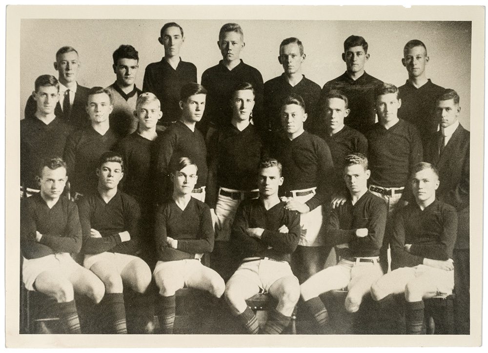 A black and white photograph of a group of young men in rugby uniforms posing for a team picture, some standing, some sitting.