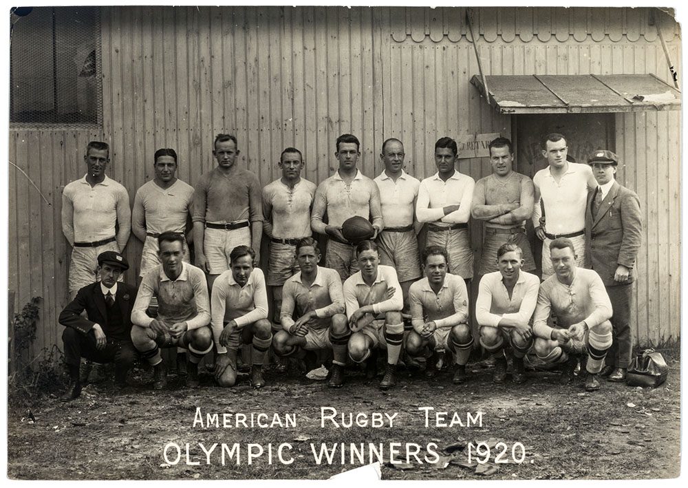 A black and white photograph of a rugby team posing in front of a wooden building in uniform. It reads "American Rugby Team Olympic Winners, 1920."