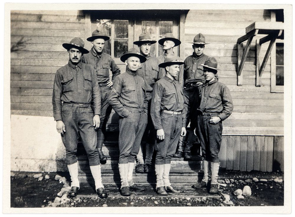 A group of eight men in early 20th century military uniforms standing on the front steps of a wooden building.
