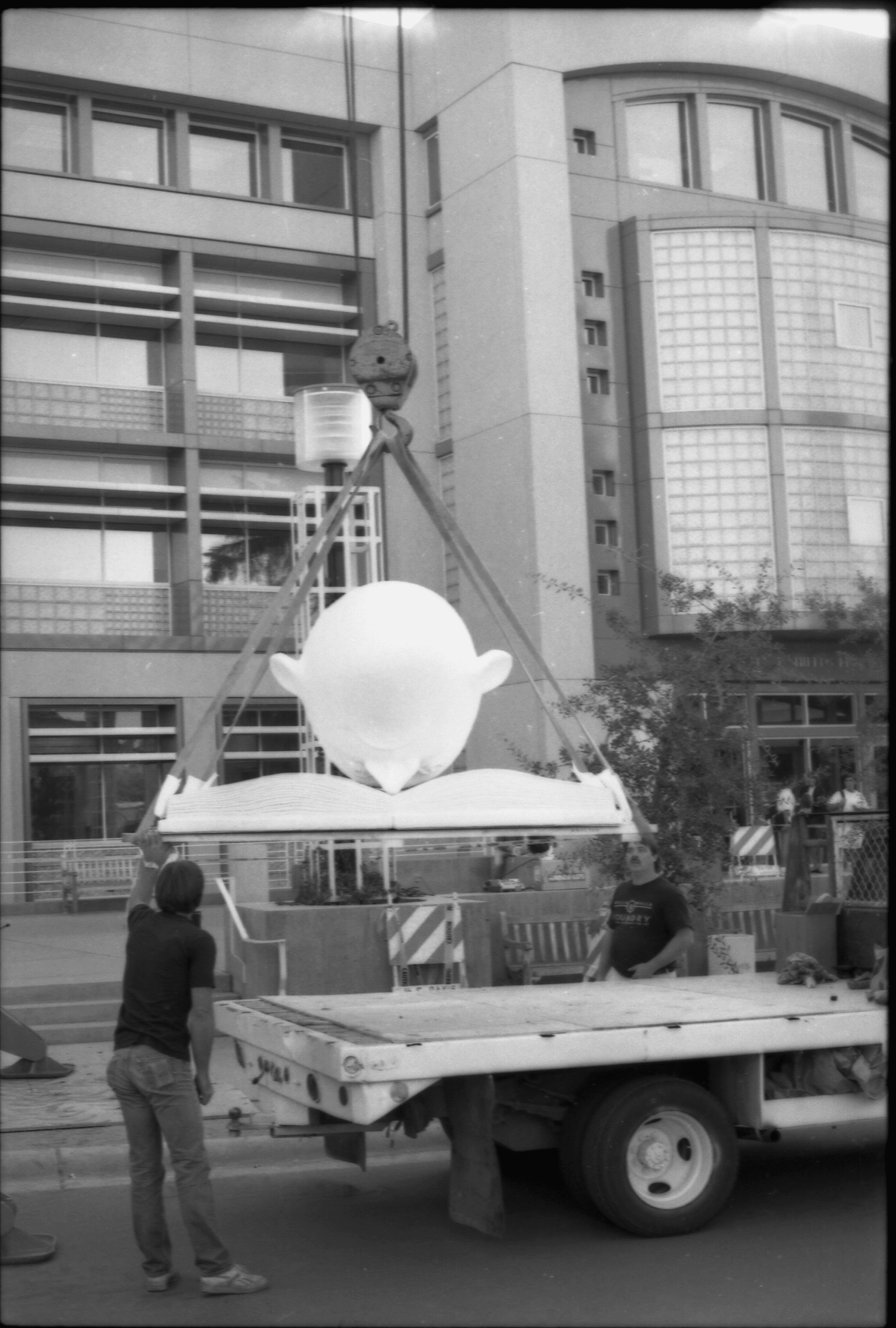 Bookhead sculpture is lifted on a crane, supervised by two workers. The entrance to Shields Library is visible in the background.