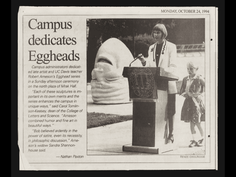 Newspaper clipping with a photo of a woman standing at a podium with a young girl beside her. Behind them is the "Eye on Mrak" Egghead sculpture.
