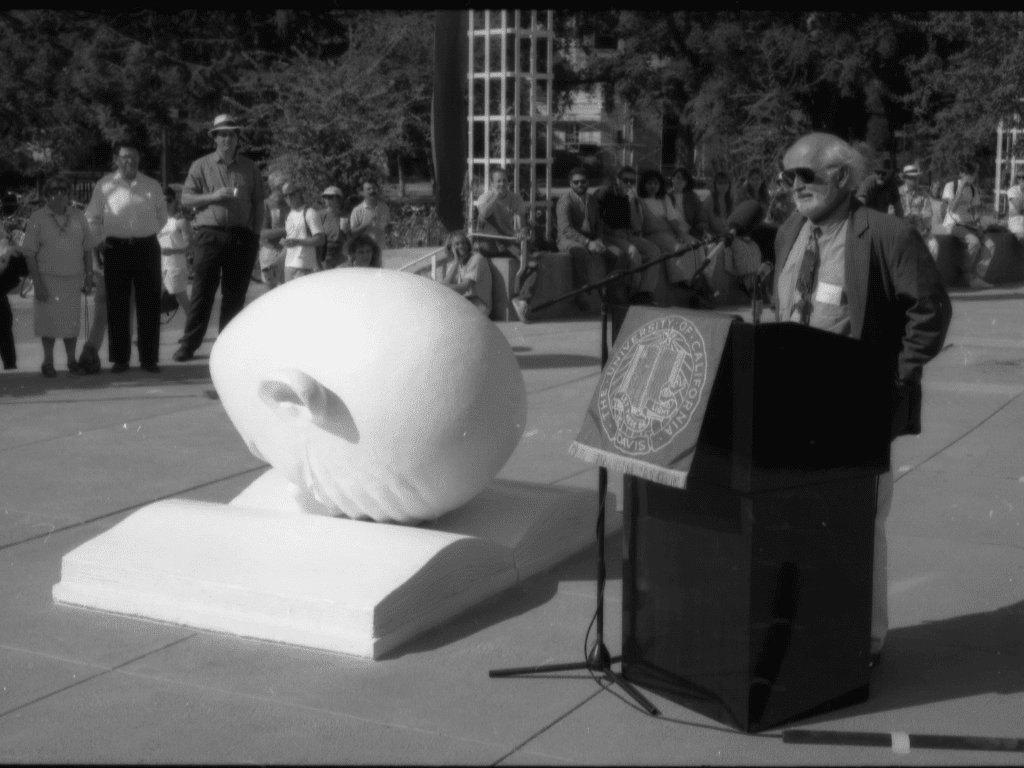 Robert Arneson stands at a podium next to the Bookhead sculpture. A row of onlookers is in the background.