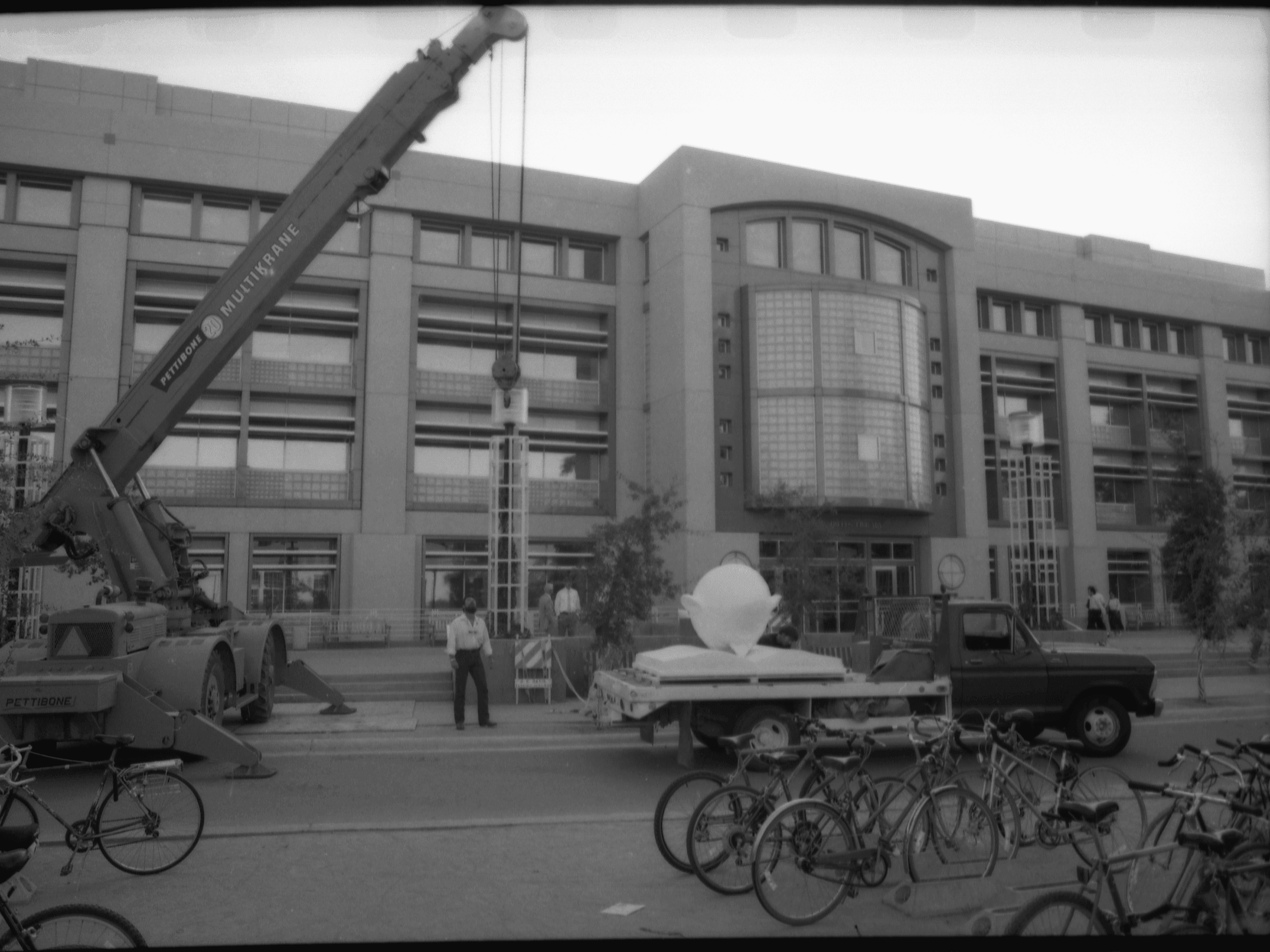 Black-and-white photo of Bookhead sculpture on the back of a flatbed truck in front of Shields Library. A crane is behind the truck.