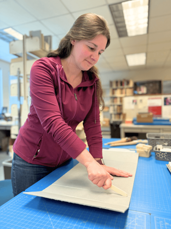 A woman with a hard, flat white strip in her hand pressing it into a piece of gray cardboard.