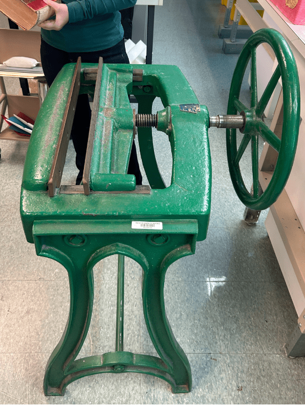 A large green mechanical device with a round wheel used to facilitate compressing text blocks and backings together.