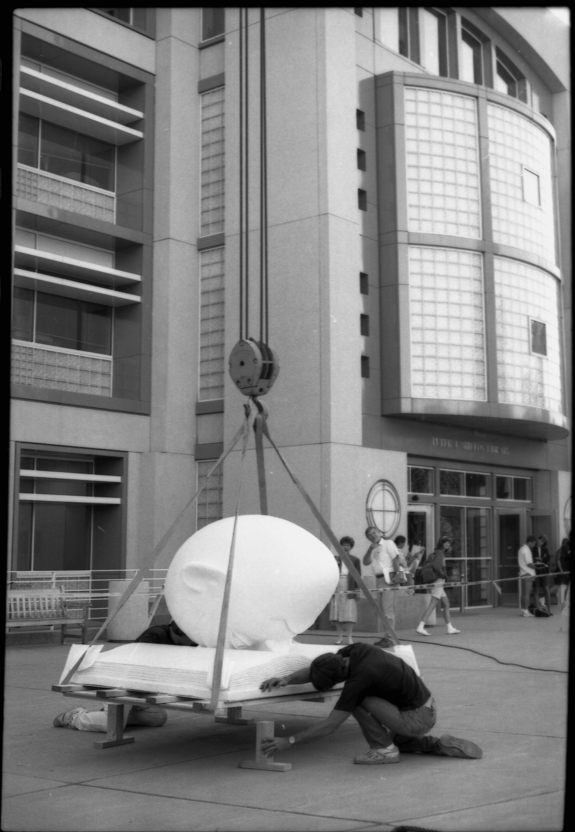 Bookhead sculpture is lowered to the ground by a crane. A worker kneels by the sculpture to align it.