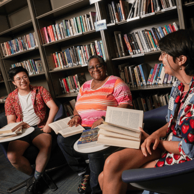 three people sitting in front of bookcases, with books in their hands and on small tables in front of them