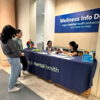 Two students sit behind a desk draped with a blue "aggie mental health" tablecloth. Flyers and a spin-the-wheel game are on top of the desk. Two other students stand in front of the desk. All four students are smiling.