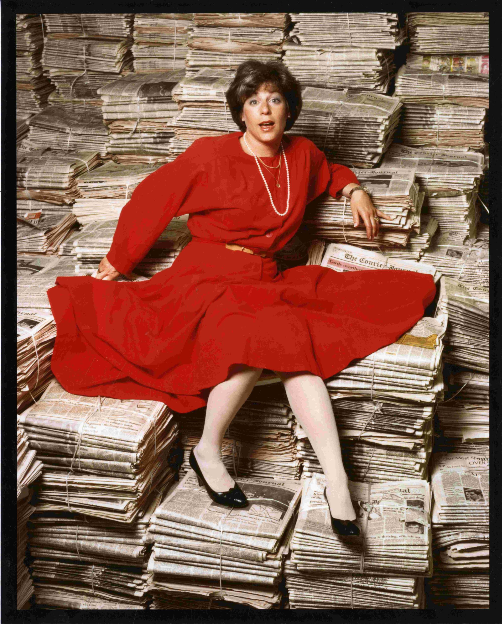 A woman wearing a red dress, pearl necklace and black high heels is seated on numerous bound stacks of newspapers. She stares at the camera with a fun, surprised expression.