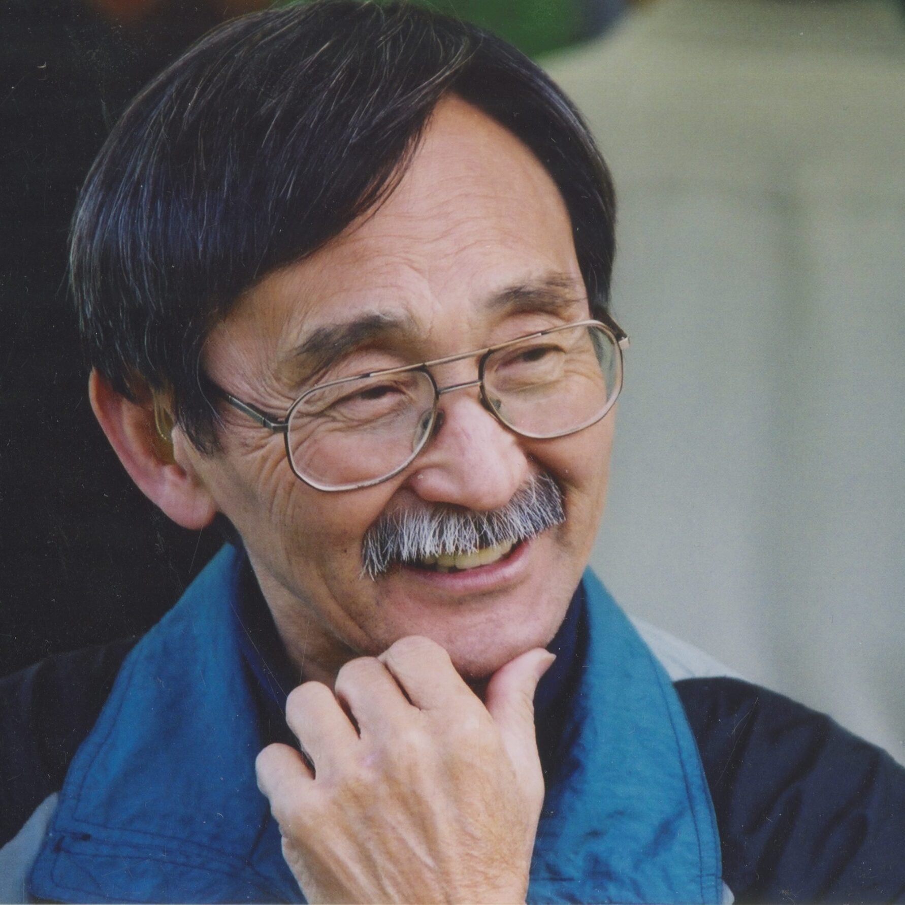 An older man with glasses smiling with a thoughtful hand to his chin.