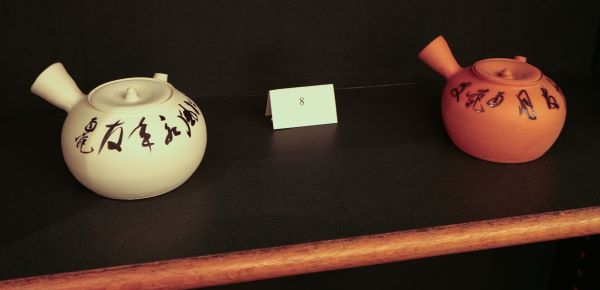 Two ceramic water kettles, decorated with Japanese script in ink, on a shelf.