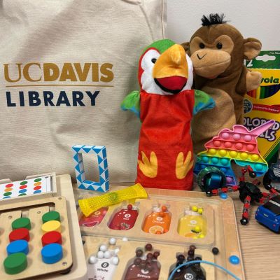 Objects arranged on a table including puzzles, puppets, a toy truck and a box of colored pencils. A UC Davis Library tote bag is in the background.