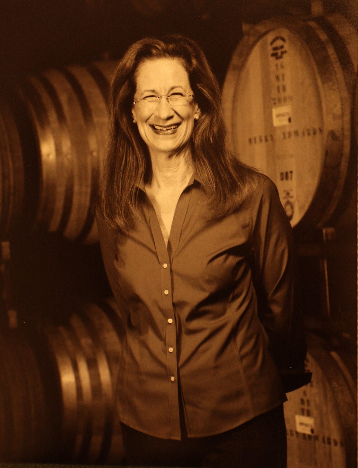 A sepia photograph of a woman with long dark hair and glasses smiling at the camera. She is standing in front of several wine barrels.
