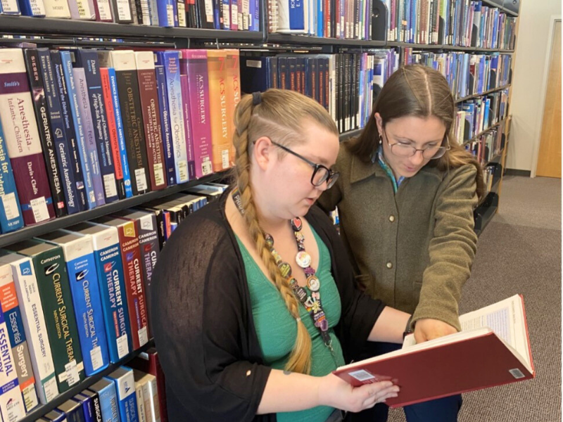 two librarians looking at a book with bookshelves behind them.