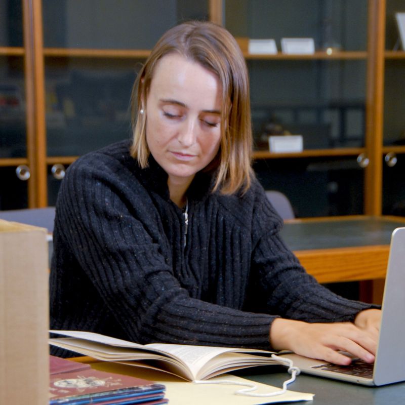 A young woman working on a laptop while simultaneously reading a booklet.