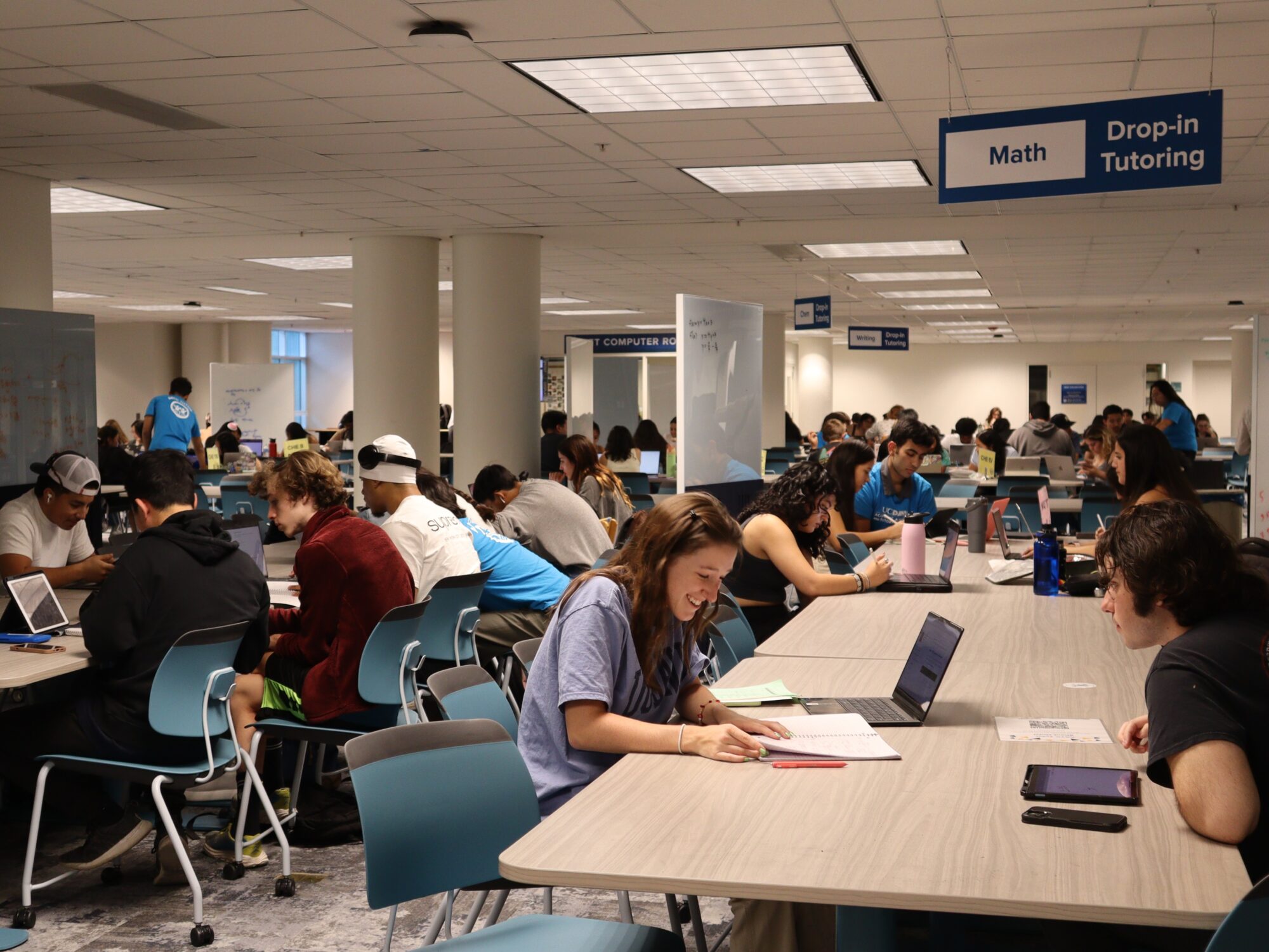 A brightly lit room filled with students studying together. A sign hanging from the ceiling reads "Math Drop-in Tutoring"