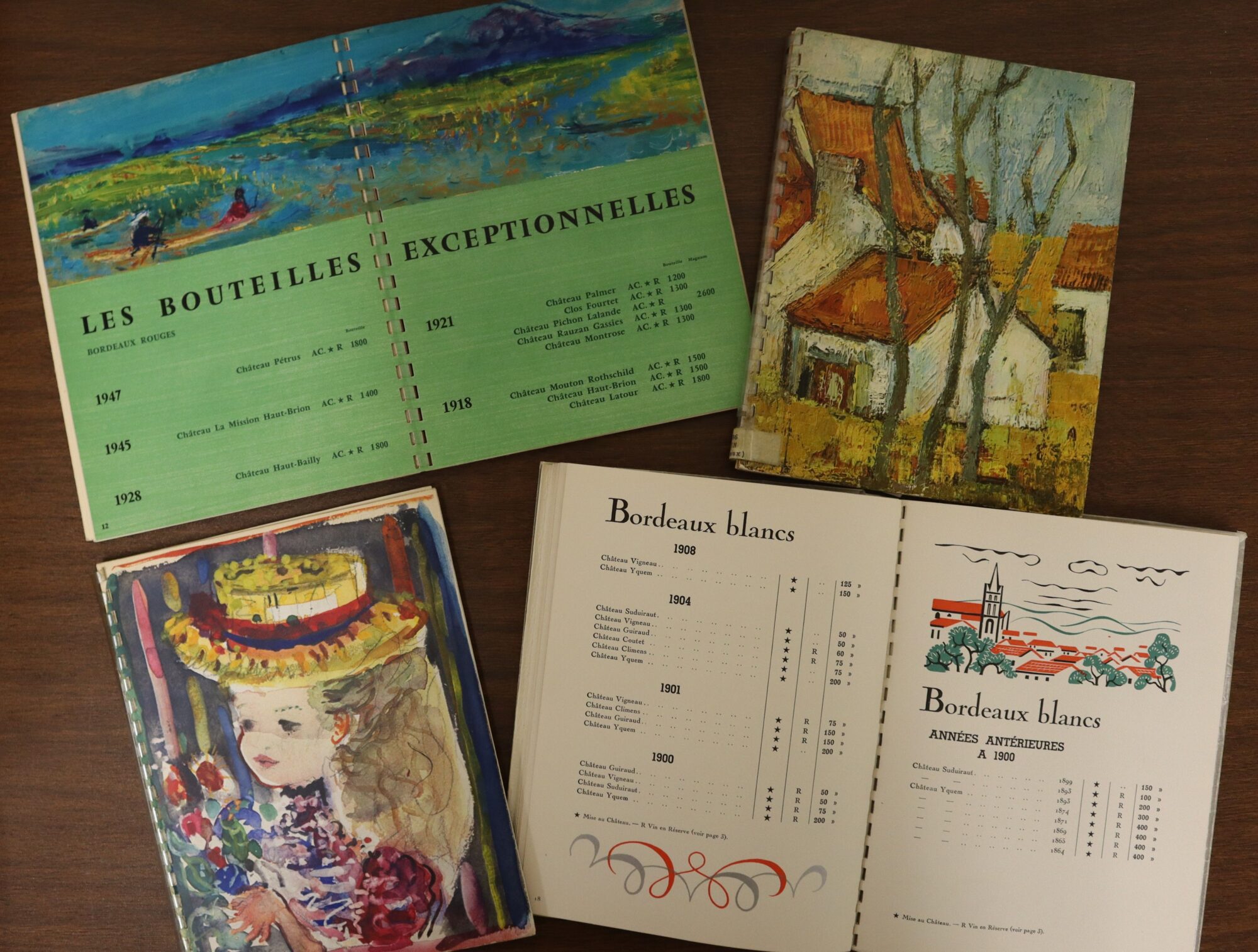 Four wine catalogs on a tabletop. Two are open to show inside pages; the other two show the paintings on their covers.