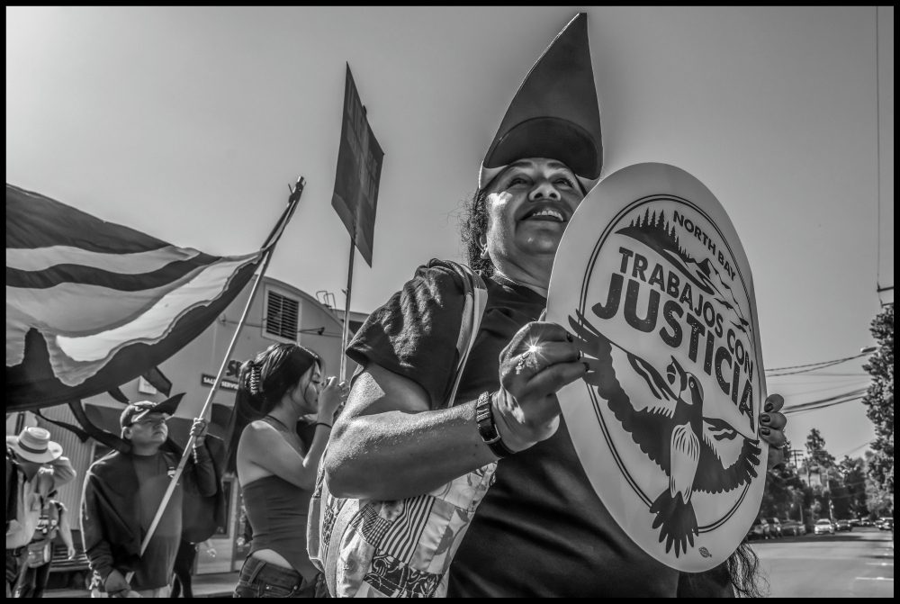A woman holds a sign that reads North Bay Trabajos con Justicia. Other protestors hold signs and flags in the background.