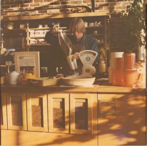 An old photograph of a blond woman behind a wooden counter pouring coffee beans from one bag to another.