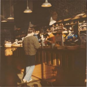 An old photograph of people, one in front of a wooden counter and two behind it. A brick wall with wooden shelves full of coffee bags is in the background.