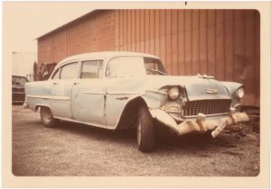 An old photograph of a sideways-facing blue car sitting in front of a brown paneled wall on concrete.