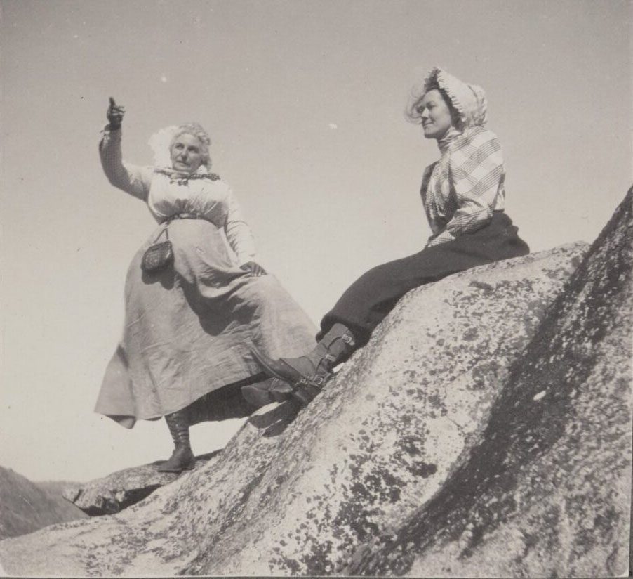 A black and white photograph of two women in 19th century dresses standing on a rocky ridge.